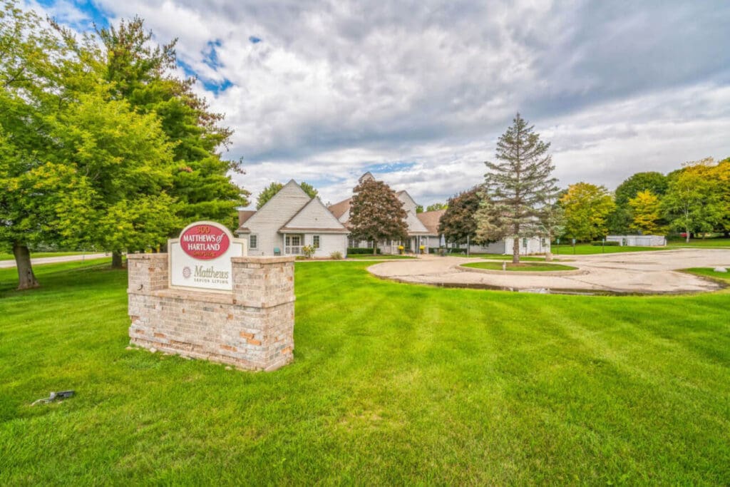 A grassy area with a stone sign for Matthews of Neenah serves as a peaceful setting reminiscent of Hartland memory care. Behind it, white buildings with gabled roofs are embraced by trees. The sky is cloudy, and a circular driveway is visible, enhancing the serene atmosphere.
