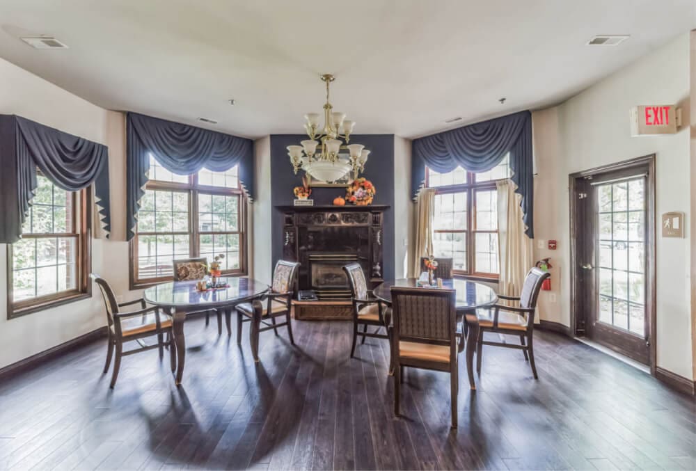 A well-lit dining room with four wooden chairs around two connected glass tables. A chandelier hangs from the ceiling. The room has large windows with blue curtains, and a decorative fireplace is centered between the windows, adorned with flower vases.