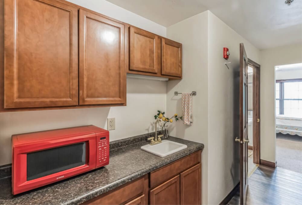 Kitchenette with wooden cabinets, a red microwave on a dark countertop, and a small sink. A towel hangs nearby with decorative flowers in a vase. A doorway leads to another room with natural light.