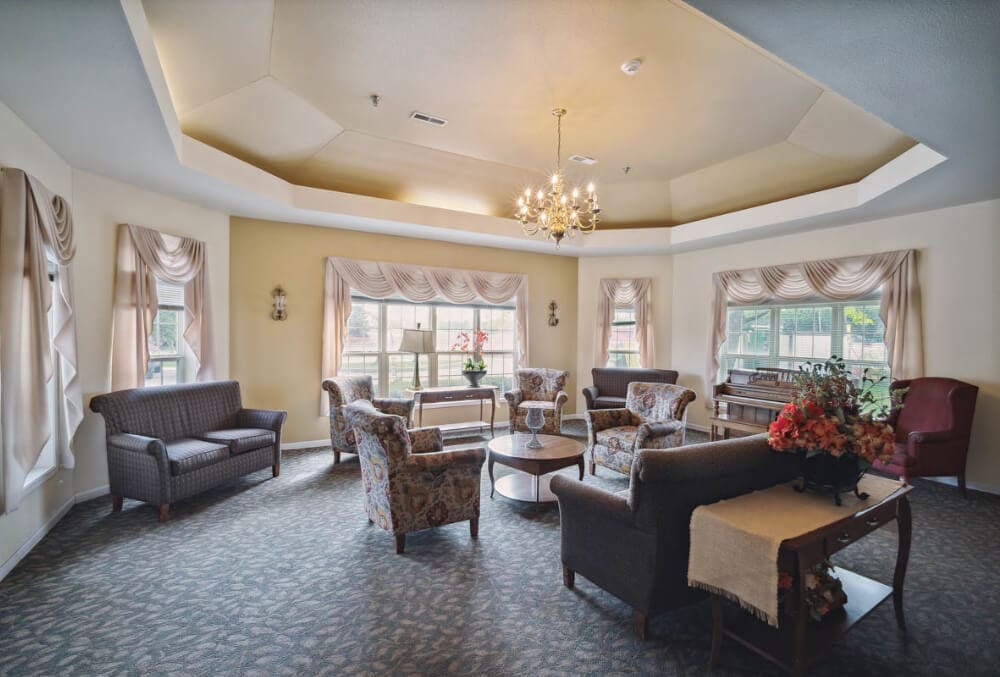 A cozy living room with a circular arrangement of upholstered chairs and sofas around a wooden coffee table. A chandelier hangs from the ceiling. Large windows with draped curtains let in natural light, and a piano is in the background.