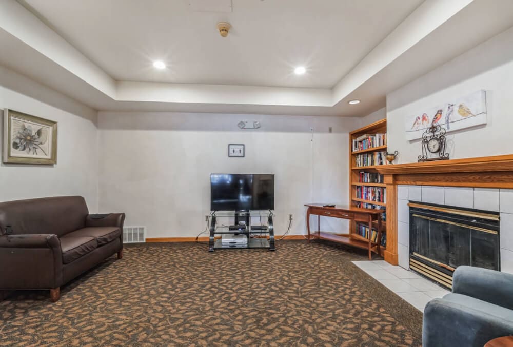 Living room with a brown sofa and a TV on a stand in front. A fireplace with a white mantle is on the right, and there are bookshelves beside it. The carpet is patterned, and the walls are adorned with framed art. Recessed lighting brightens the space.