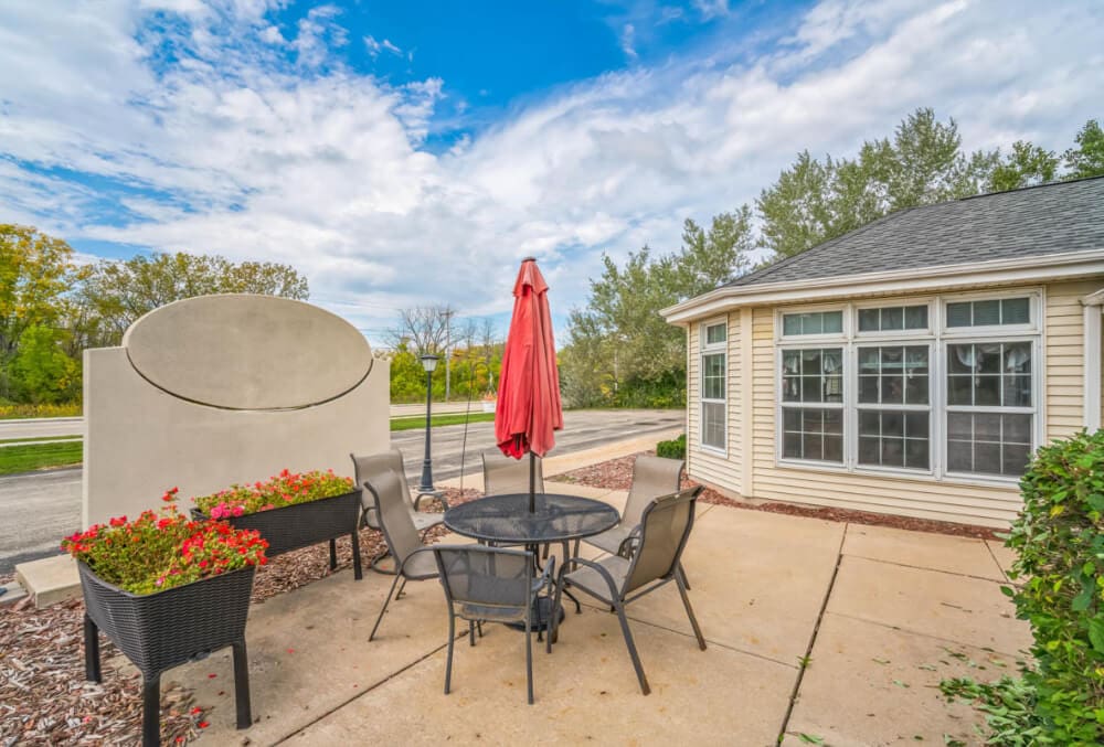Patio scene with a round black table and four chairs under a red umbrella. Nearby are two planters with red flowers. A small building with large windows is on the right. The background features a cloudy sky and green trees.