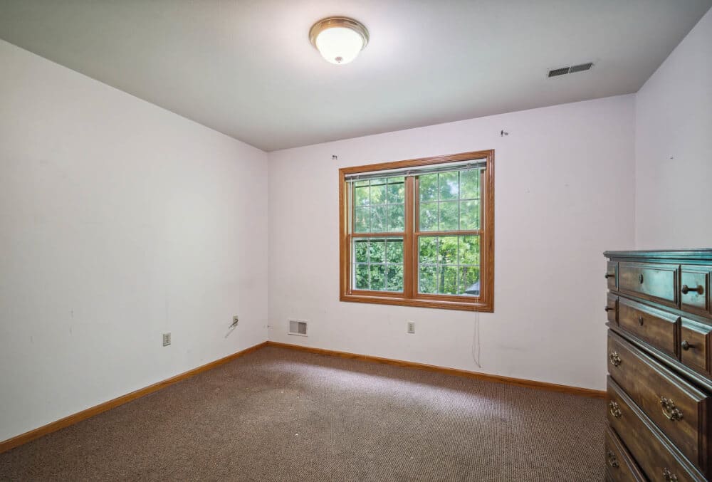 An empty room with white walls and a beige carpet. A wooden dresser is on the right, and a large window with a wooden frame is centered on the back wall, showing greenery outside. A ceiling light is on, and the room appears well-lit.