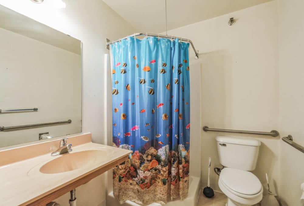 Bathroom featuring a light-colored countertop with a sink, a toilet, and a shower with a vibrant aquatic-themed curtain depicting various fish and coral. The walls are light, and there are silver grab bars near the toilet and shower.