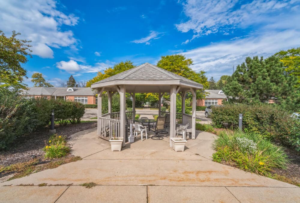 A wooden gazebo with a gray shingle roof sits on a concrete path surrounded by greenery. Inside are several chairs and a table. In the background, there are trees, shrubs, and residential buildings under a blue, partly cloudy sky.