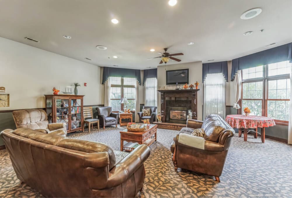 A cozy living room with brown leather sofas arranged around a coffee table. A fireplace with a TV above is centered on the far wall. Large windows with curtains and a patterned carpet complete the rooms decor.