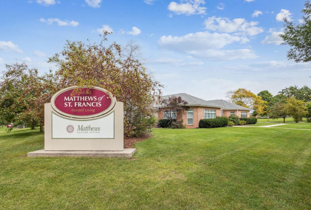 Sign reading Matthews of St. Francis Assisted Living on a grassy lawn with trees and a single-story brick building in the background under a partly cloudy sky.