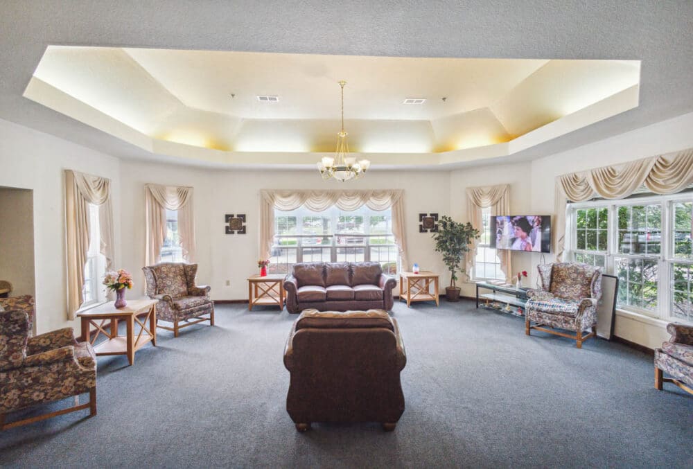A spacious living room features a central chandelier, a dark brown sofa and armchair, patterned chairs, and a television on the wall. Large windows with cream curtains surround the room, providing natural light. Two small side tables hold lamps.