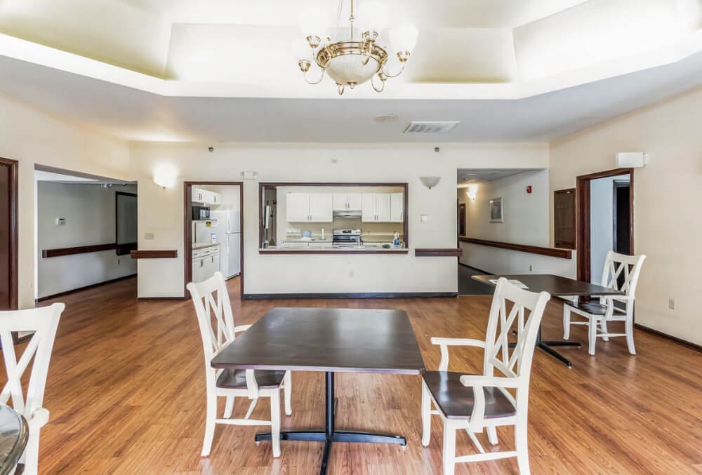 A bright dining room with wooden flooring, featuring a chandelier, white walls, and a series of tables and chairs. A counter and open view into a kitchen area are visible in the background. Natural light illuminates the space.