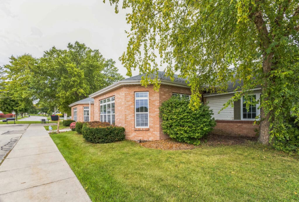 A single-story red brick house with a gabled roof is surrounded by green grass and trees. Large windows face the front, and theres a concrete sidewalk running alongside the property. The sky is overcast.