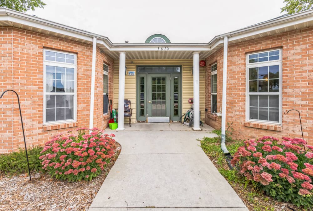 A brick building with a central entrance, featuring a green door and white trim. Two large windows flank the entrance. Pink flowering bushes line the pathway leading to the door. A small, covered porch with a white roof is above the doorway.