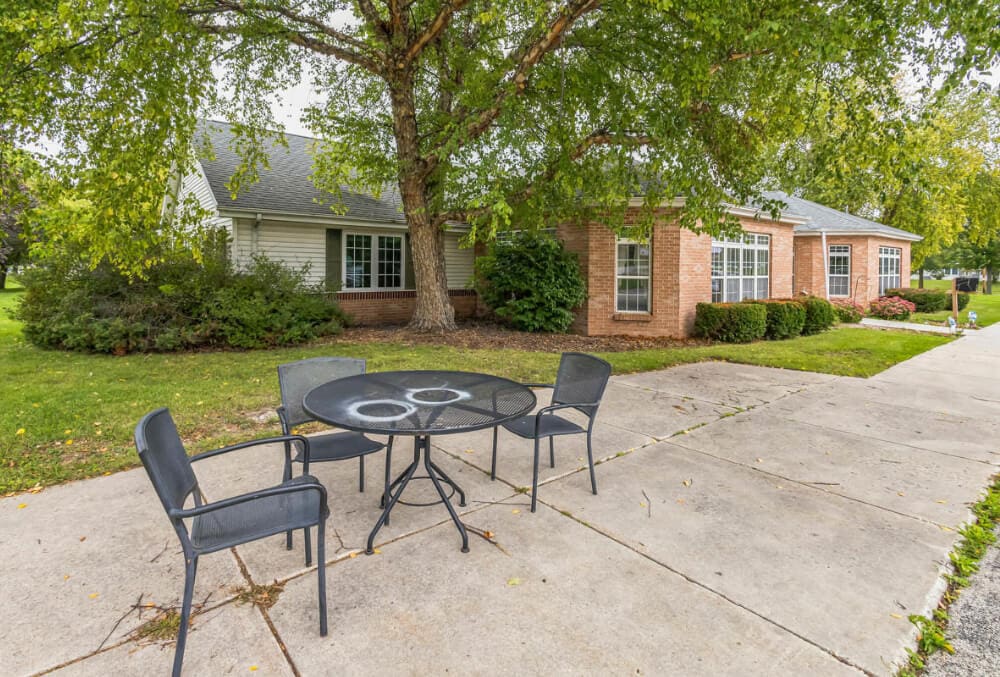 A patio with a black metal table and four chairs is set on a wide concrete pathway. Nearby, a brick and white paneled building is partially visible, surrounded by green grass and trees with abundant leaves.