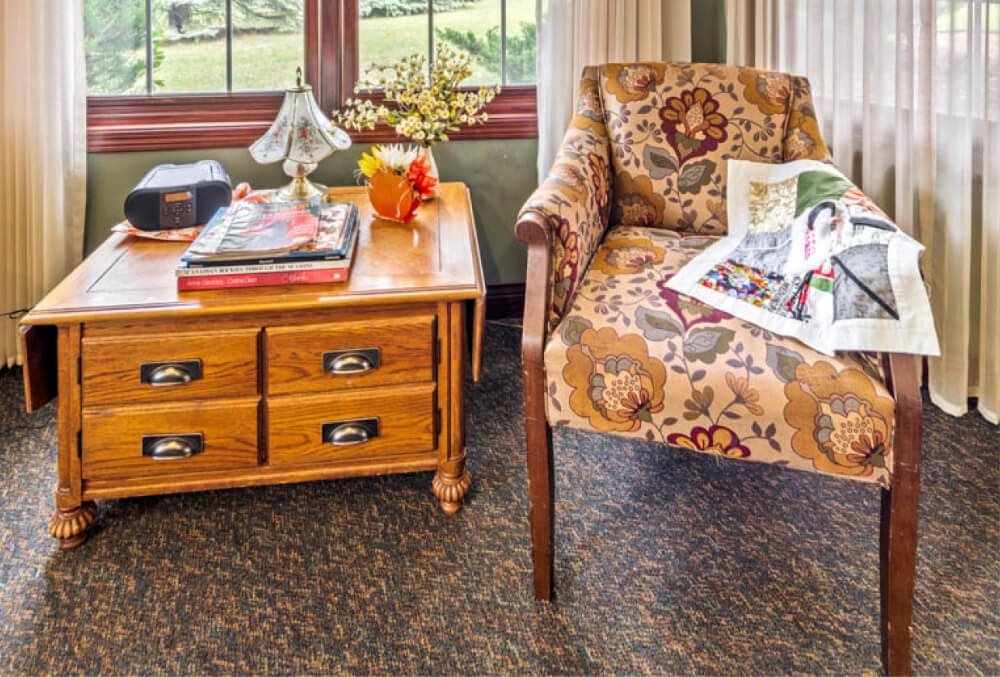 A cozy room with a patterned armchair and a magazine draped over it. A wooden side table beside the chair holds a lamp, a radio, and books. Large windows with sheer curtains let in natural light, illuminating the floral carpet.