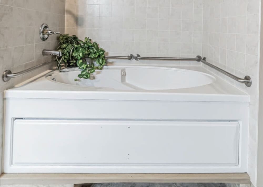 A white bathtub with a surrounding handrail is installed against a tiled wall. A green leafy plant is placed on the tubs edge, with its leaves cascading downwards. The overall setting appears clean and minimalistic.