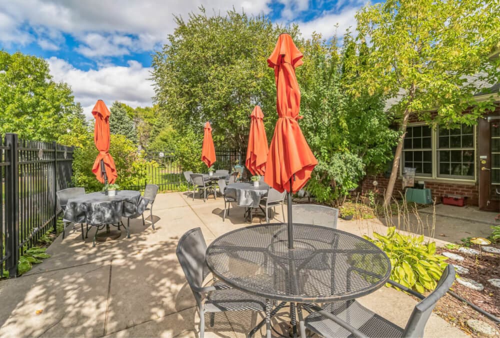 Outdoor patio with round metal tables and chairs, each table covered with a gray cloth. Red umbrellas are closed above the tables. The area is surrounded by green trees and shrubs, and theres a building on the right. Sky is partly cloudy.