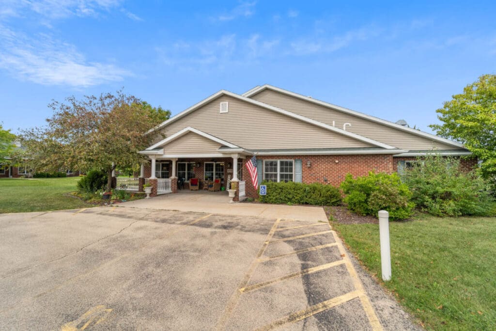 A single-story brick building with a covered entrance and an American flag stands proudly in Neenah. The area, part of the senior care facility, features a parking space with yellow markings, lush green lawns, tall trees, and a clear blue sky.