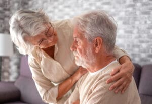 An older woman with gray hair and glasses lovingly embraces an older man with a beard, who is sitting on a couch. The woman is gently supporting him, and they both appear thoughtful in a softly lit room.