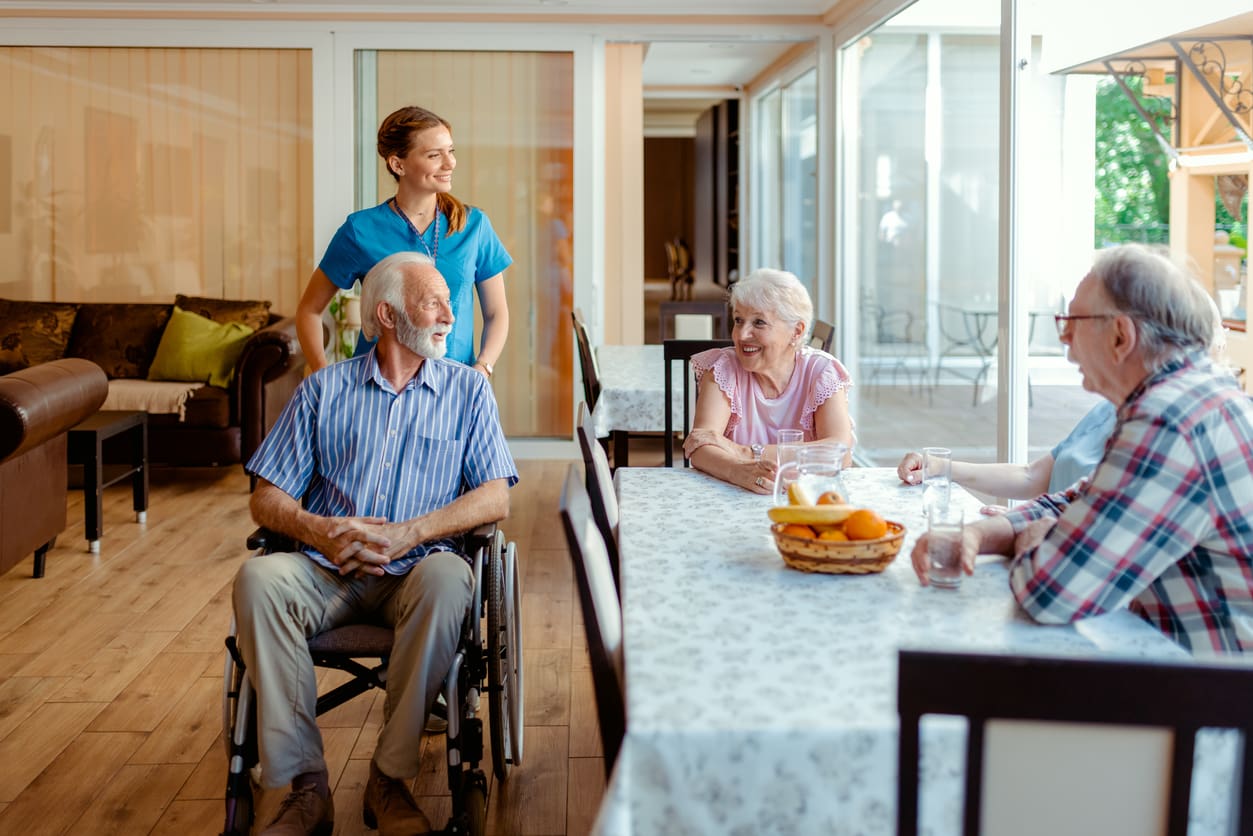A healthcare worker in blue scrubs smiles while standing behind an older man in a wheelchair. Three older adults sit at a table with a fruit basket, chatting and smiling. The room is bright with large windows.